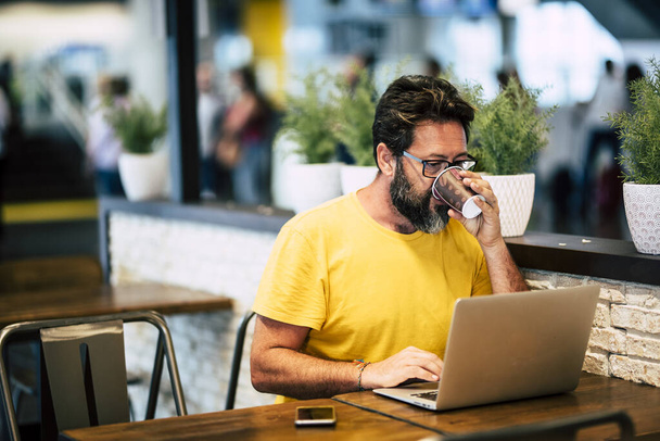 Красавчик-хипстер с бородой пьет кофе в коворкинге и работает с ноутбуком и бесплатным беспроводным доступом в Интернет  - Фото, изображение