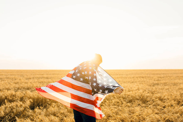 attractive man holding the flag of the United States against a blue sky on a clear sunny day, view from the back, close-up. National holiday concept - Photo, Image