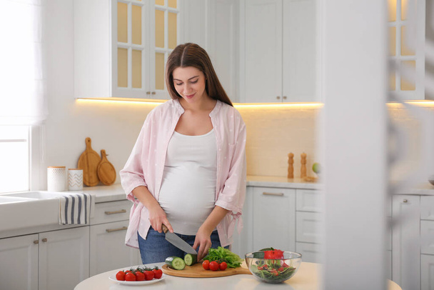 Young pregnant woman preparing vegetable salad at table in kitchen. Healthy eating - Photo, Image