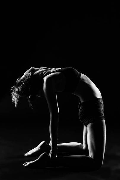 A grayscale shot of a woman in sportswear doing yoga in a studio with a dark background - Фото, зображення
