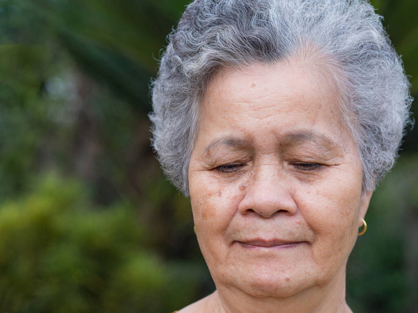 Close-up van het gezicht van een oudere vrouw die glimlacht en neerkijkt terwijl ze buiten staat. Ruimte voor tekst. Begrip ouderen en gezondheidszorg. - Foto, afbeelding
