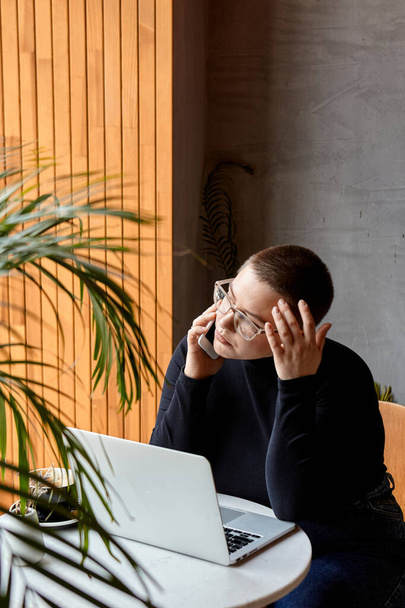 Rapariga com cabelo curto e nariz piercing senta-se em um café. Uma mulher está a falar ao telefone e a trabalhar num tablet. O conceito de trabalho freelancer e remoto. - Foto, Imagem