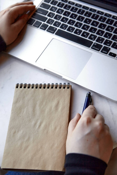 Uma jovem está sentada em um café, trabalhando em um laptop e fazendo anotações em um caderno. Fechar - acima da foto de mãos. O conceito de trabalho freelancer e remoto ou treinamento. - Foto, Imagem