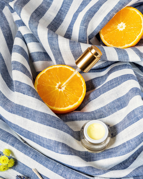 The concept of natural skin care products and cosmetics made from organic ingredients.Orange halves with a cosmetic pipette and a jar of cream on striped textile background. Citrus cream for the face - Foto, Bild