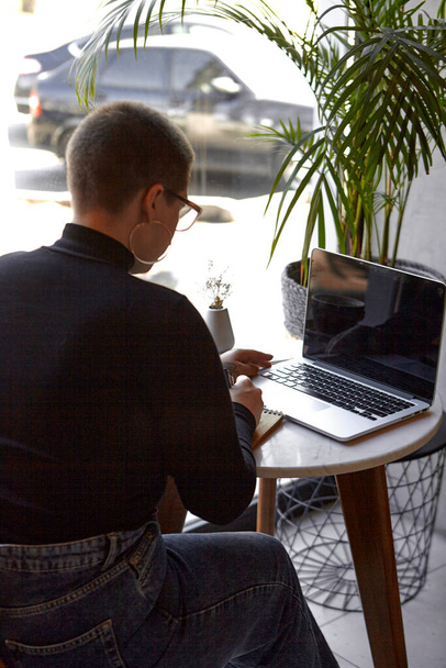 Młoda dziewczyna w okularach, krótkich włosach i kolczyku w nosie siedzi w kawiarni i pracuje nad laptopem. Koncepcja freelancingu i zdalnej pracy lub szkolenia. - Zdjęcie, obraz