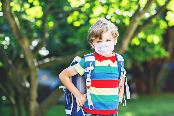 Happy little kid boy with medical mask and backpack or satchel. Schoolkid on way to school. Healthy child outdoors. Back to school after quarantine time from corona pandemic disease lockdown - Φωτογραφία, εικόνα