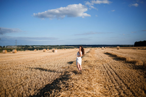 Портрет молодой кудрявой женщины на пшеничном поле, где косят пшеницу и стоят снопы, наслаждаясь природой. Природа. солнечные лучи - Фото, изображение