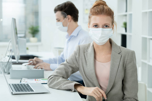 Young elegant businesswoman in protective mask looking at you on background of her male co-worker analyzing online data - Foto, immagini
