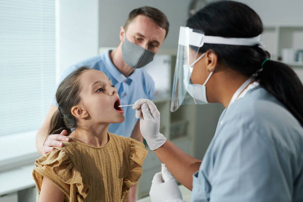 Kleines Mädchen mit offenem Mund sitzt vor einer Krankenschwester gemischter Rassen in Uniform und Schutzkleidung und testet sie auf Covid mit Wattestäbchen - Foto, Bild