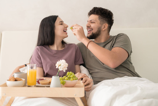 junger, fröhlicher Mann, der seiner Frau Trauben in den Mund steckt, während beide unter einer weißen Decke im Doppelbett sitzen und das Frühstück genießen - Foto, Bild
