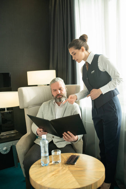 Young female worker of hotel in uniform bending over mature male guest with menu choosing food for breakfast while sitting in armchair - Photo, Image