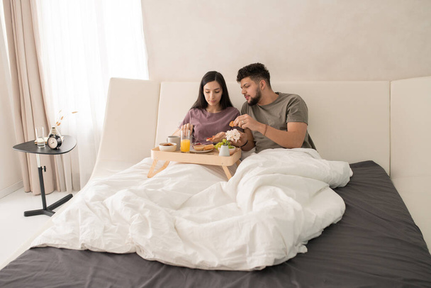 Młoda czuła para w t-shirtach siedzi w łóżku i je smaczne śniadanie, podczas gdy samica rozrzuca dżem na plasterku chleba pszennego - Zdjęcie, obraz