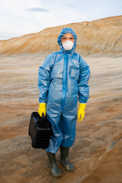 Young female researcher in protective workwear holding case with samples while standing on polluted soil in abandoned area against hill and sky - Photo, Image