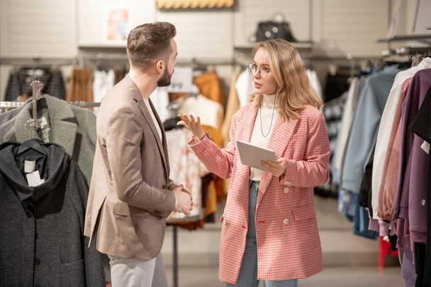Junge Stylistin mit digitalem Tablet im Gespräch mit dem Kunden im Bekleidungsgeschäft im Einkaufszentrum - Foto, Bild