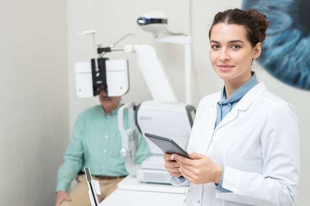 Heureux jeune ophtalmologiste femelle dans whitecoat vous regardant tout en utilisant la tablette sur fond de patient ayant sa vue vérifiée - Photo, image
