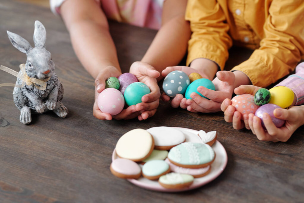 Manos de tres niños pequeños interculturales en ropa casual sosteniendo huevos de Pascua pintados entre galletas y conejo de juguete mientras está sentado junto a la mesa - Foto, Imagen