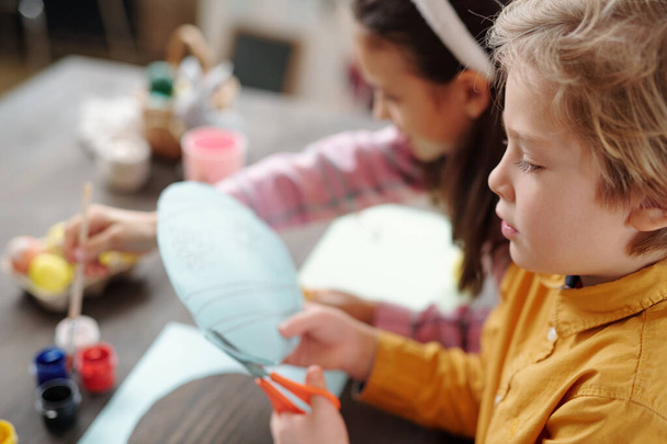 niedliche blonde kleine Junge schneidet Papier Osterkarte, während sie am Holztisch auf dem Hintergrund seines Freundes Gemälde Bild oder Eier - Foto, Bild