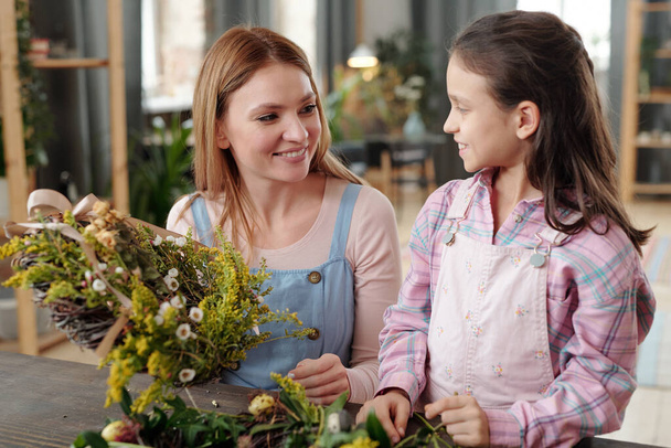 Happy young female in casualwear looking at her little daughter with toothy smile while both standing by table and preparing Easter wreath - Foto, Bild