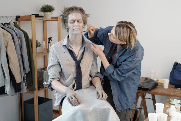 Çağdaş genç iş adamı veya aktörün yüzüne ve boynuna zombi makyajı yapan sarışın yüzlü bir sanatçı. - Fotoğraf, Görsel
