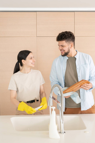 Ευτυχισμένος νεαρός άνδρας και γυναίκα κοιτάζονται με χαμόγελα ενώ τα θηλυκά πλένουν τα πιάτα και ο σύζυγός της σκουπίζει τον πίνακα κοπής - Φωτογραφία, εικόνα
