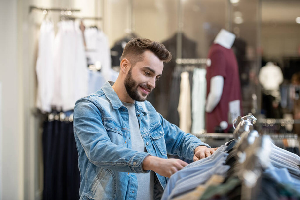 Young bearded man choosing new clothes on the rack in shopping mall - Photo, Image