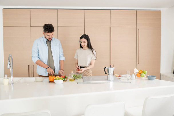 Onnellinen nuori pari casualwear seisoo keittiön pöydän ääressä ja ruoanlaitto kasvissalaatti koostuu kurkut, tomaatit ja paprika - Valokuva, kuva