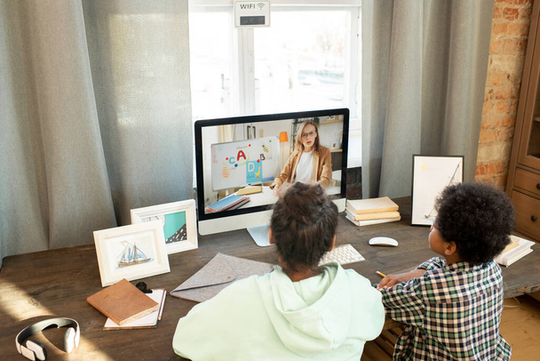 Dva smíšení sourozenci v neformálním oblečení sedící u stolu před počítačovou obrazovkou a poslouchající jejich učitelské vysvětlení během online lekce - Fotografie, Obrázek