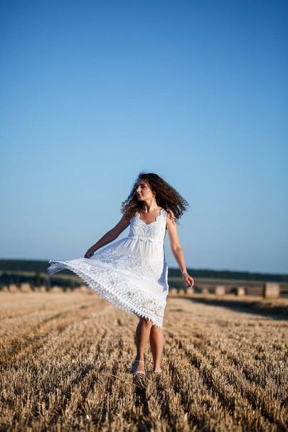 A young beautiful woman in a white summer dress stands on a mown wheat field with huge sheaves of hay, enjoying nature. Nature in the village. Selective focus - Foto, Imagen