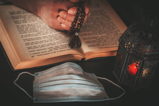 Hands with prayer beads over the holy Muslim book of the Koran, dark background with glowing Arabic lantern.y and a medical mask. Celebrating Holy month Ramadan in quarantine. Epidemic, coronavirus pandemic or covid-19 concept - Photo, Image
