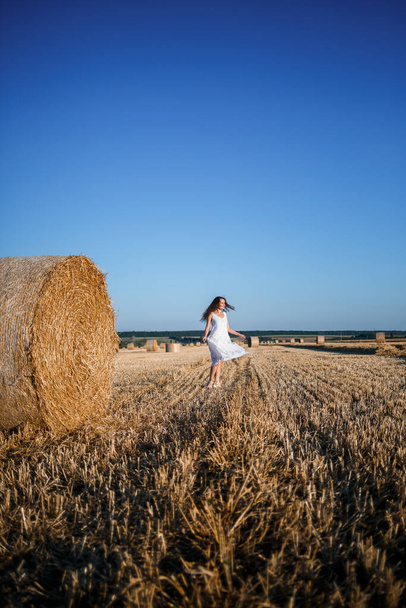 Nuori kaunis nainen valkoisessa kesämekossa seisoo niitetyllä vehnäpellolla, jolla on valtavat heinät, nauttien luonnosta. Kylän luonto. Valikoiva painopiste - Valokuva, kuva