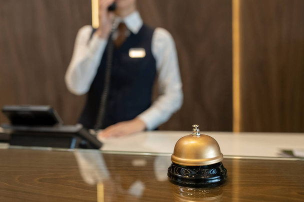 Bell on wooden reception counter against female receptionist consulting clients on the phone while working in contemporary luxurious hotel - Foto, immagini