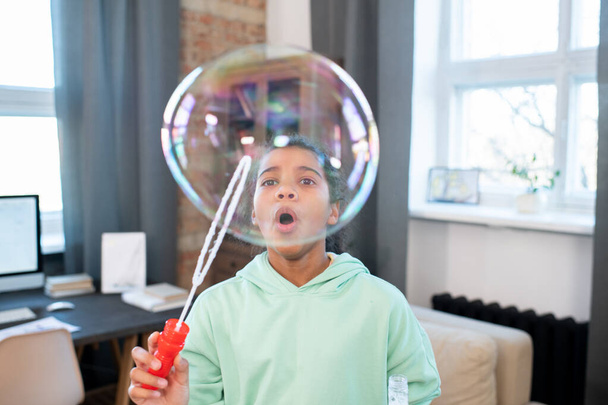 Cute girl of elementary age in casualwear blowing huge soap bubble while standing in front of camera and having fun in home environment - Photo, Image