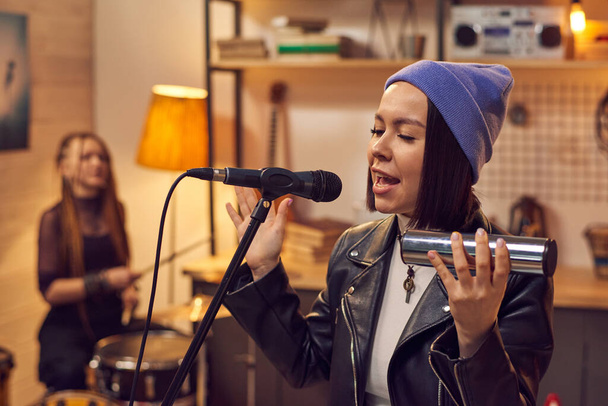 Young beautiful singer in stylish hat playing musical instrument and singing in microphone in music studio - Photo, Image