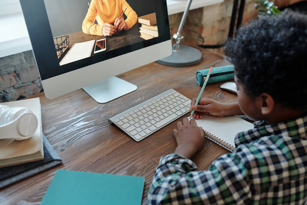 Malý chlapec sedí u stolu před monitorem počítače během on-line lekce, poslouchá svého učitele na obrazovce a dělá si poznámky do sešitu - Fotografie, Obrázek
