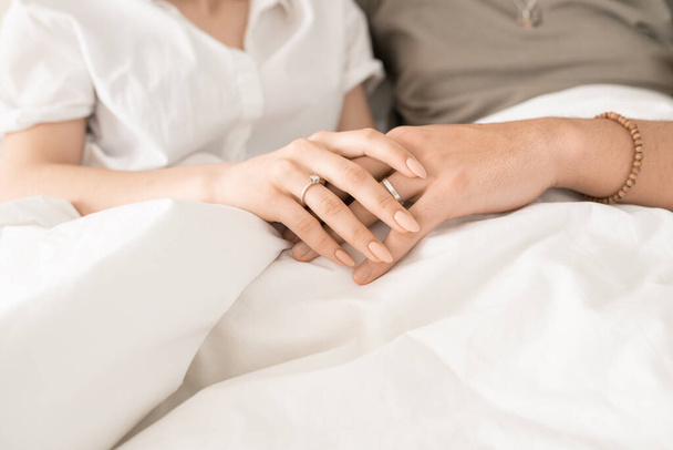 Hände von jungen verliebten Eheleuten, die es sich morgens nach dem Schlafen oder beim Fernsehen unter einer weißen Decke im Bett gemütlich machen - Foto, Bild