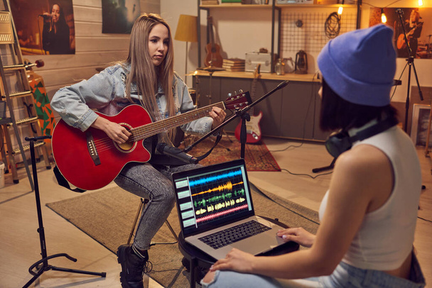 Two young women recording a song together in the studio using laptop and playing guitar - Φωτογραφία, εικόνα