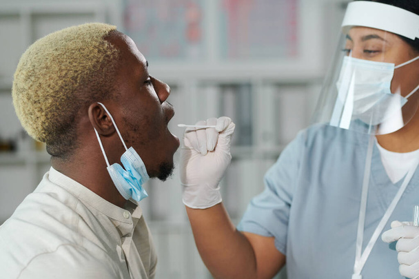 Mladá smíšené rasy žena klinik v ochranném pracovním oděvu stojí před africkým mužem s otevřenou pusou, zatímco ho testuje na covid - Fotografie, Obrázek
