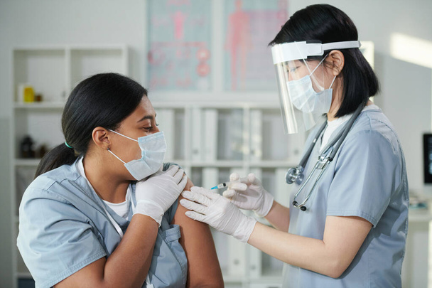 Korumalı iş elbisesinde çalışan genç Asyalı hemşire kliniklerde covid aşısı sırasında melez bayan meslektaşına iğne yapıyor. - Fotoğraf, Görsel