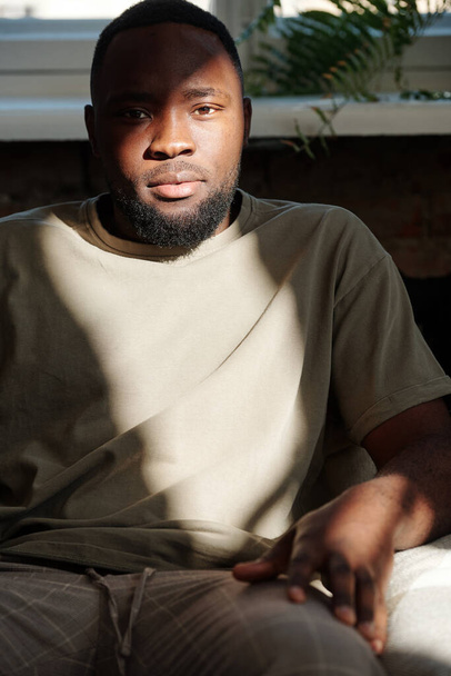 Young restful bearded man of African ethnicity looking at you while sitting in armchair in front of camera against windowsill of living-room - Photo, Image