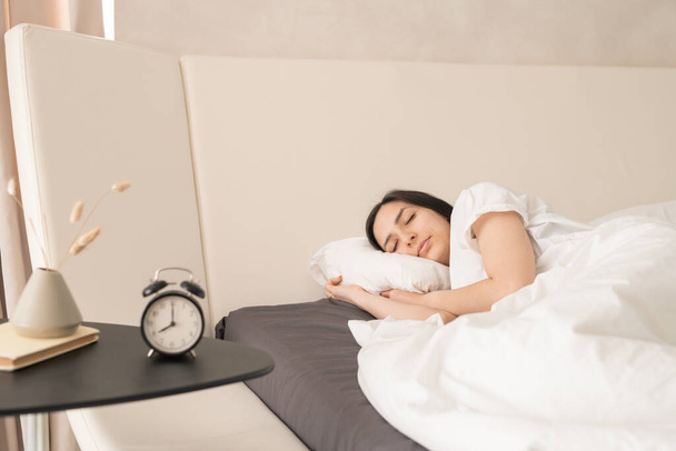 Mujer hermosa joven que duerme tranquilamente en una cama doble grande y cómoda delante de una pequeña mesa con reloj despertador, jarrón y libro - Foto, Imagen