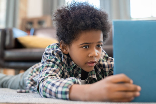Netter Grundschüler mit digitalem Tablet liegt auf dem Boden vor schwarzer Ledercouch im Wohnzimmer und schaut neugierige Online-Videos - Foto, Bild