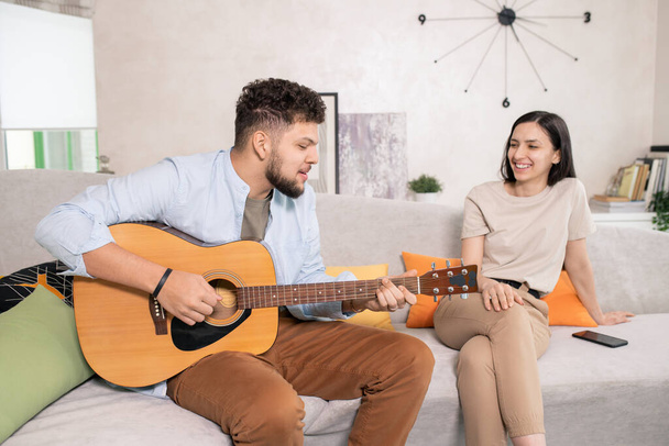 Щаслива молода жінка з зубною посмішкою сидить на дивані у вітальні і дивиться на чоловіка, який грає на гітарі і співає для неї
 - Фото, зображення