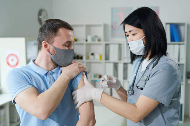 Clinicienne ou infirmière d'origine asiatique en uniforme et masque protecteur faisant injection à un jeune homme assis devant elle dans un cabinet médical - Photo, image