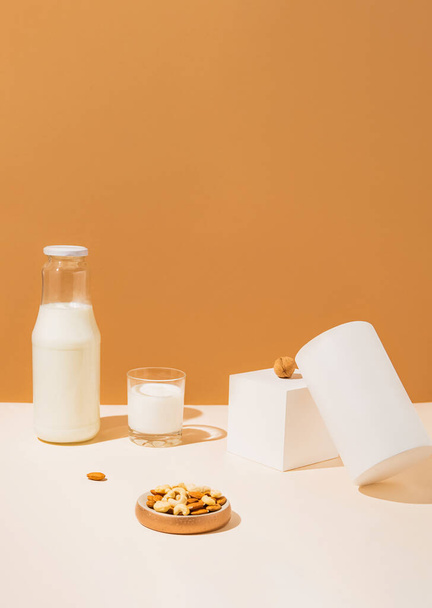 Glass and a bottle with vegan milk from various nuts with white geometric figures on sunlit background. Dairy and lactose free substitute drink. Healthy food ingredient concept. Trendy colors. - Photo, image