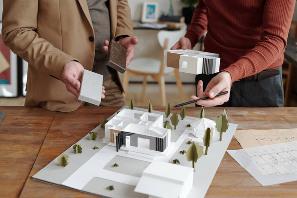 Handen van jonge mannelijke architect wijzend op model van nieuw huis en die van zijn volwassen collega die geschikt blok voor de bouw kiest - Foto, afbeelding