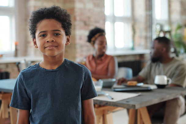 Roztomilý malý chlapec afrického etnika v tmavě modrém tričku stojící před kamerou proti jeho rodičům sedícím u kuchyňského stolu při snídani - Fotografie, Obrázek