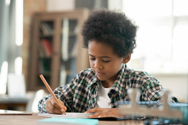 Cute serious schoolboy of African ethnicity making notes in copybook while doing homework by table in living-room against computer - Photo, Image