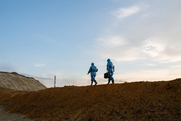Figuren zweier zeitgenössischer Ökologen in Schutzanzügen, die sich während der Untersuchung auf einem Hügel mit schmutzigem Boden gegen bewölkten Himmel bewegen - Foto, Bild