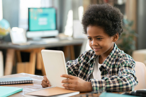 Cute elementary schoolboy of African ethnicity sitting by table in front of camera in living-room and watching online educational video - Photo, Image