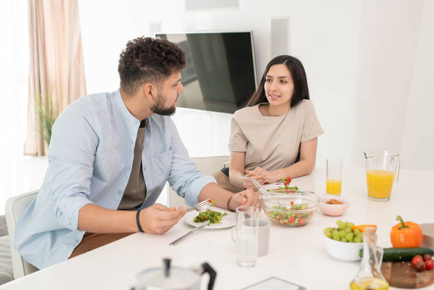 Νεαρή καστανή γυναίκα κοιτάζει τον άντρα της κατά τη διάρκεια της συνομιλίας με το πρωινό, ενώ και οι δύο έχουν selfmade σαλάτα λαχανικών και χυμό πορτοκαλιού - Φωτογραφία, εικόνα